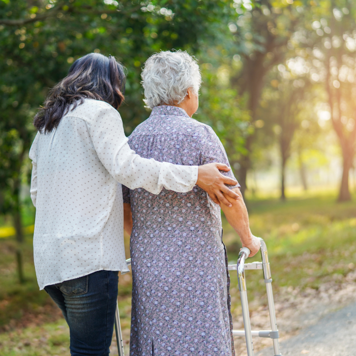Caregiver walking beside an older adult using a walker outdoors.
